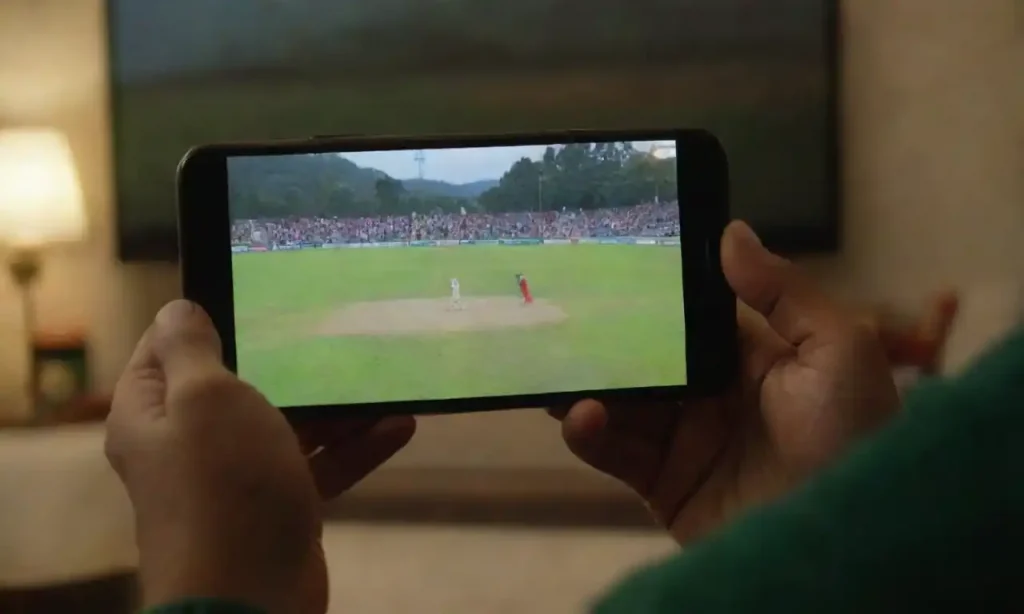 A person watching a live cricket match on a mobile device in a cozy setting.