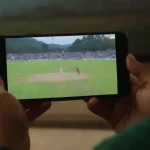 A person watching a live cricket match on a mobile device in a cozy setting.